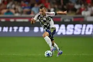 A soccer player in a black and white kit dribbles the ball during a match, with a cheering crowd in the background.