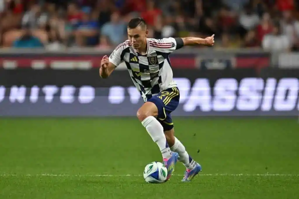 A soccer player in a black and white kit dribbles the ball during a match, with a cheering crowd in the background.
