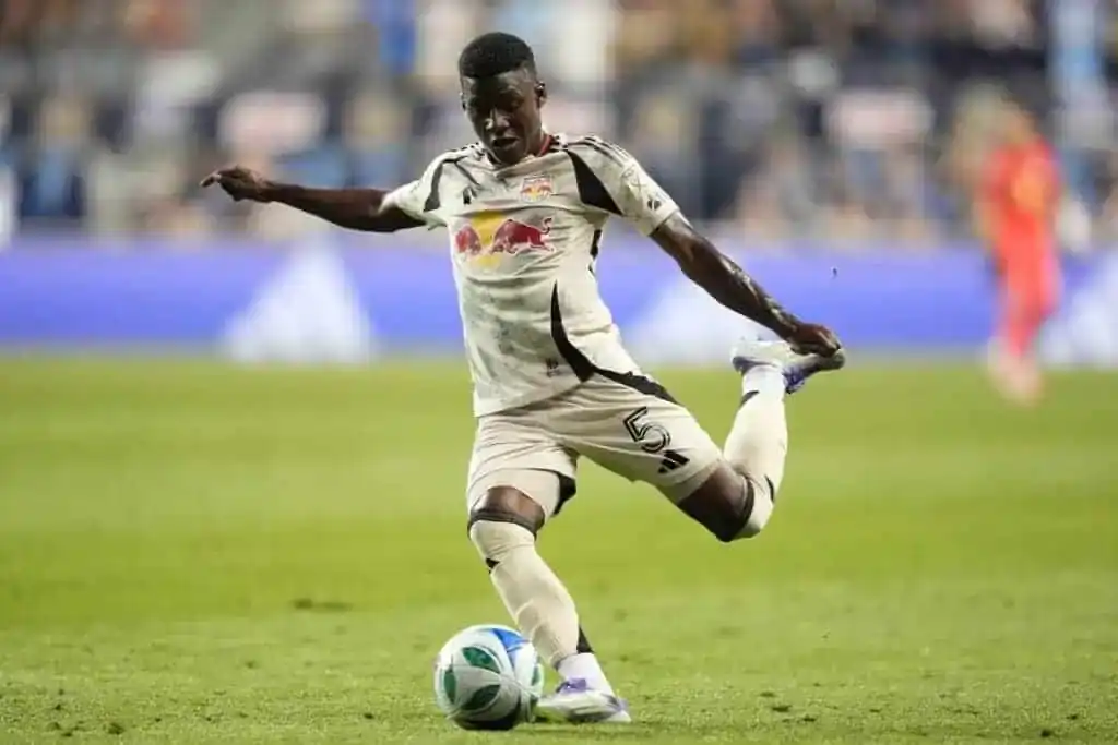 A soccer player in a light-colored jersey and shorts takes a powerful shot at the ball on a green field, with fans in the background.
