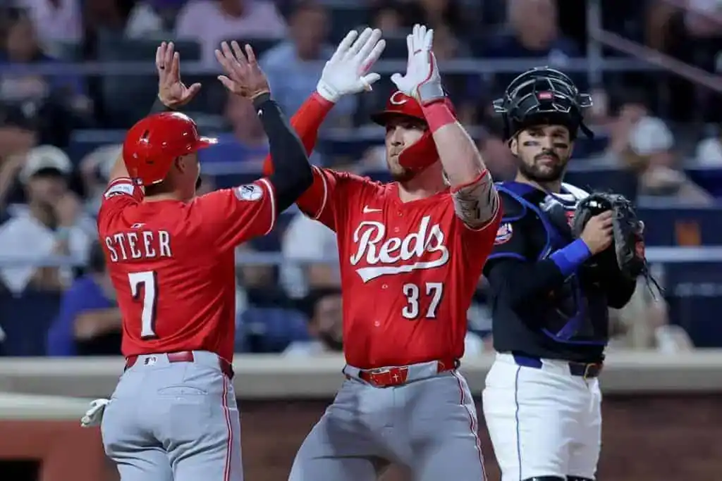 Two Cincinnati Reds players in red jerseys high-five to celebrate at home plate.