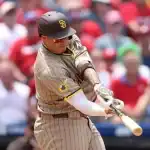 A baseball player in a striped brown and yellow uniform swings a bat at a pitch during a game, with a cheering crowd in the background.