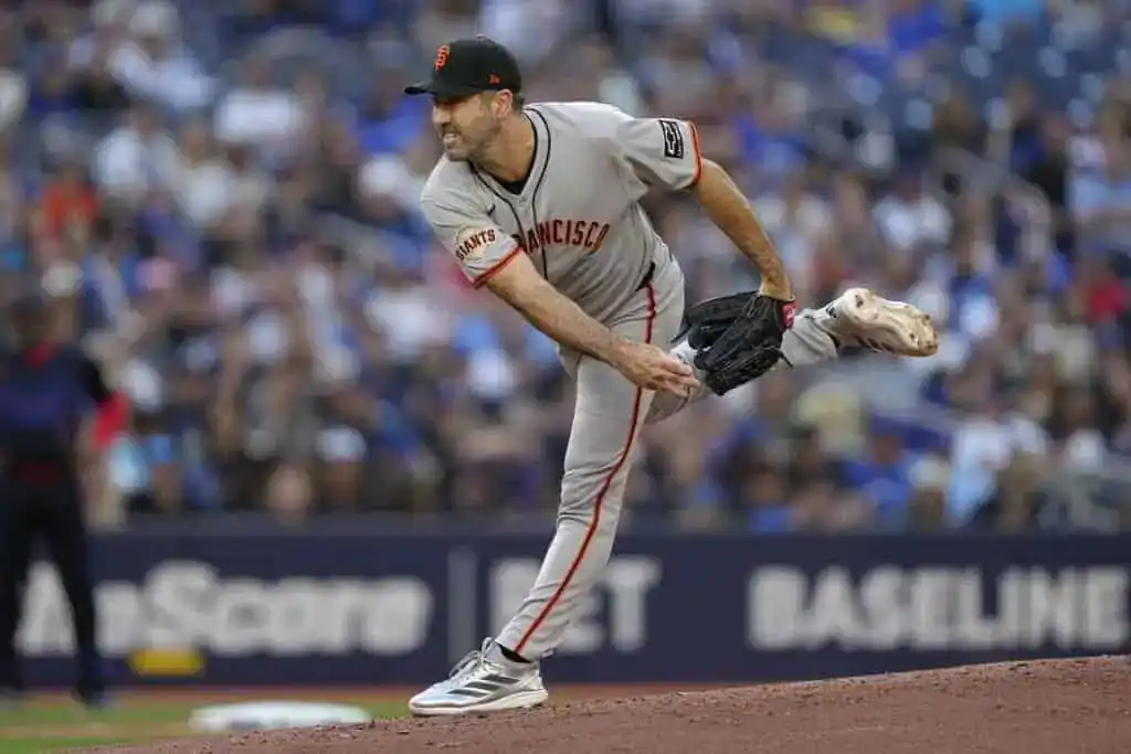 A pitcher in a gray San Francisco Giants uniform strides forward on the mound, preparing to throw a pitch against a blurred crowd background.