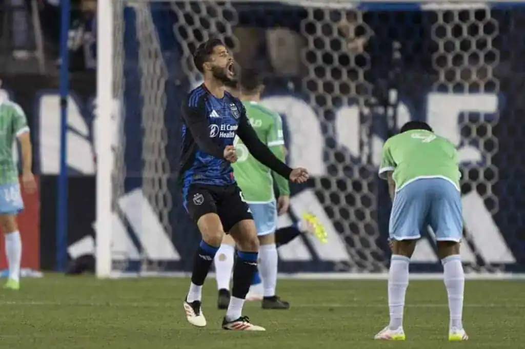A soccer match in progress, featuring players in blue and green uniforms, with a goal net in the background. Celebratory and focused actions are captured.