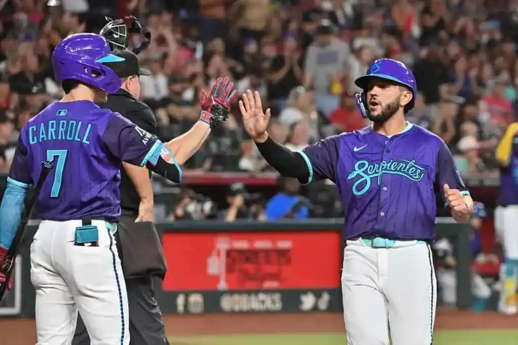 Two Arizona Diamondbacks players in purple "Serpientes" City Connect jerseys high-five on the baseball field.