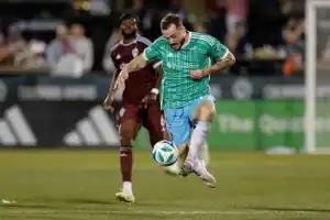 A soccer player in a green and blue jersey skillfully dribbles a ball on the field, with another player in maroon nearby.
