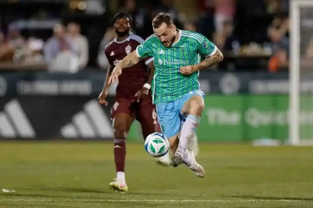 A soccer player in a green and blue jersey skillfully dribbles a ball on the field, with another player in maroon nearby.