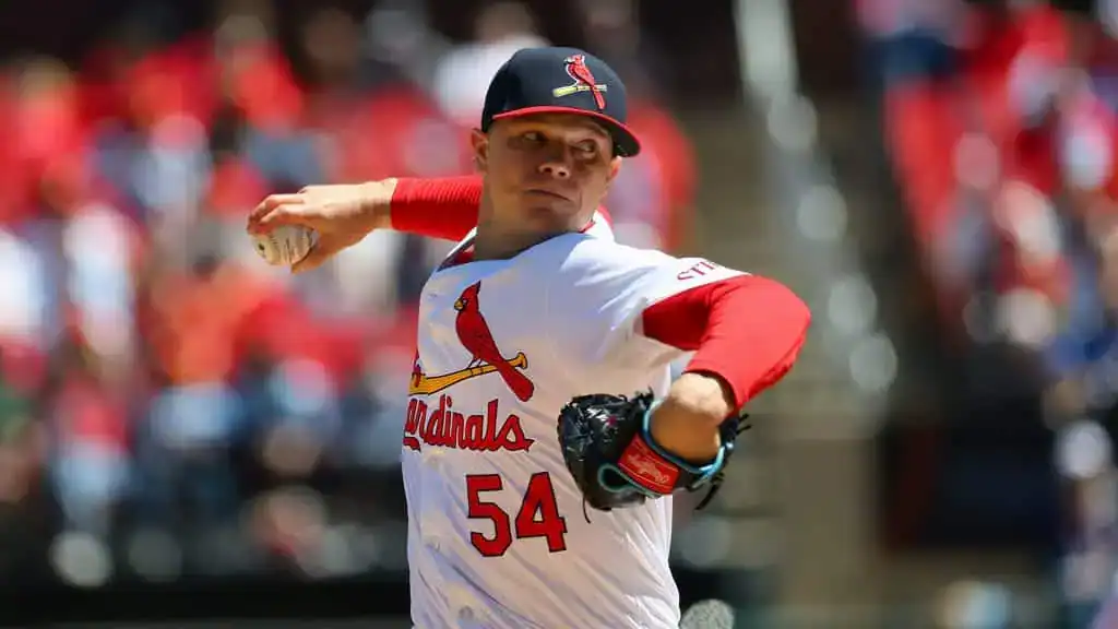 A St. Louis Cardinals player in a white jersey with red sleeves throws a pitch on a sunny day, with fans blurred in the background.