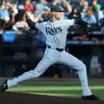 A baseball player in a Rays uniform winds up to pitch, with a packed stadium of fans in the background.