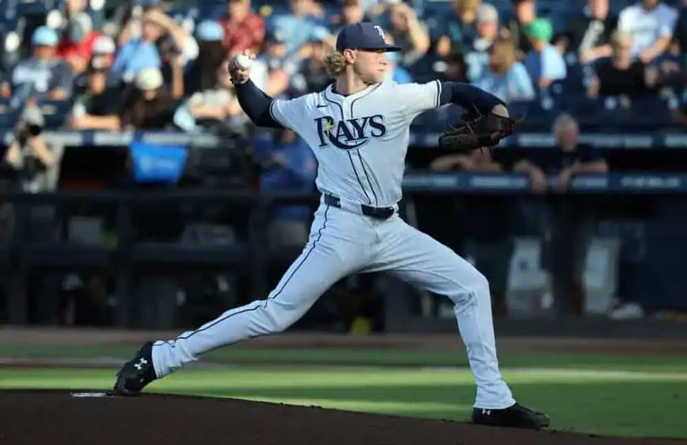 A baseball player in a Rays uniform winds up to pitch, with a packed stadium of fans in the background.