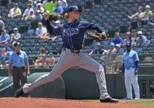 A pitcher in a dark blue Rays uniform winds up to throw during a sunny baseball game, with spectators in the background.