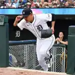A baseball player wearing a white uniform with a "D" logo runs out of the dugout toward the field, with fans watching in the background.