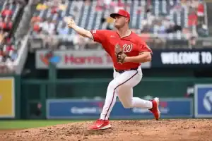 A baseball pitcher in a red jersey delivers a pitch from the mound, with fans watching in the background at a stadium.