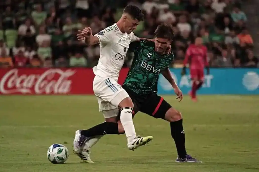 Two soccer players compete for the ball on a lush green field during a game, surrounded by a crowd cheering in the background.