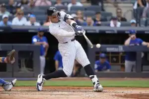 A baseball player in a pinstriped uniform swings at a pitch, with the ball just leaving the bat amid a lively baseball stadium scene.