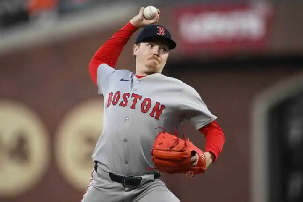 A Boston Red Sox player in gray and red uniform winds up to pitch a baseball on a baseball field.