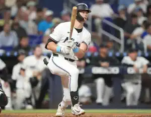 A baseball player in a Miami Marlins uniform prepares to swing at a pitch during a game, with the crowd visible in the background.