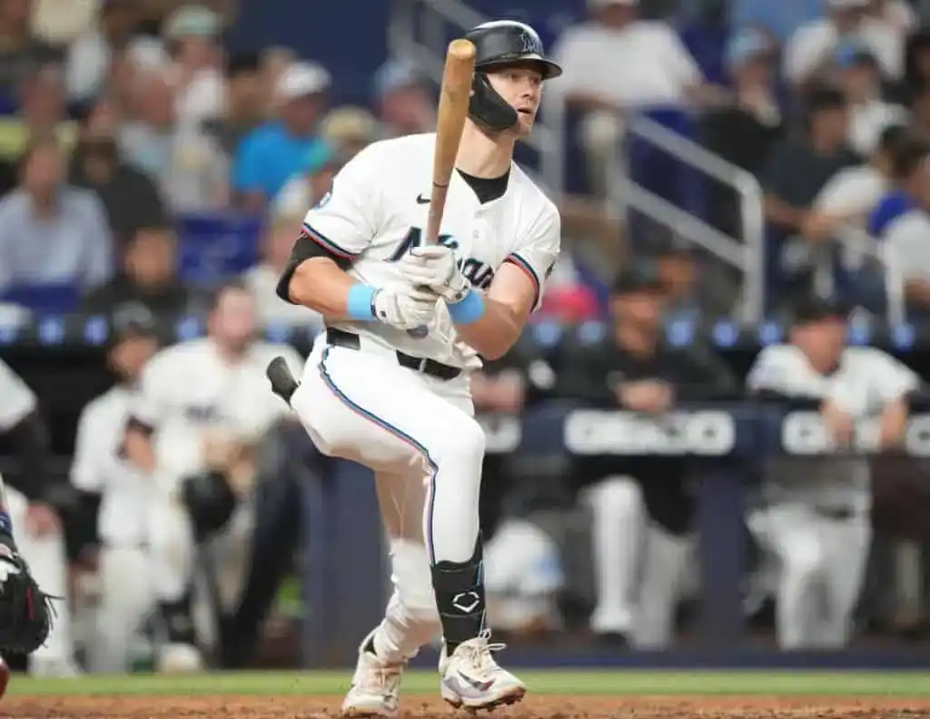 A baseball player in a Miami Marlins uniform prepares to swing at a pitch during a game, with the crowd visible in the background.