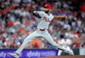 A Philadelphia Phillies pitcher throws a pitch on the baseball mound, wearing a gray uniform and a red cap, with fans in the background.