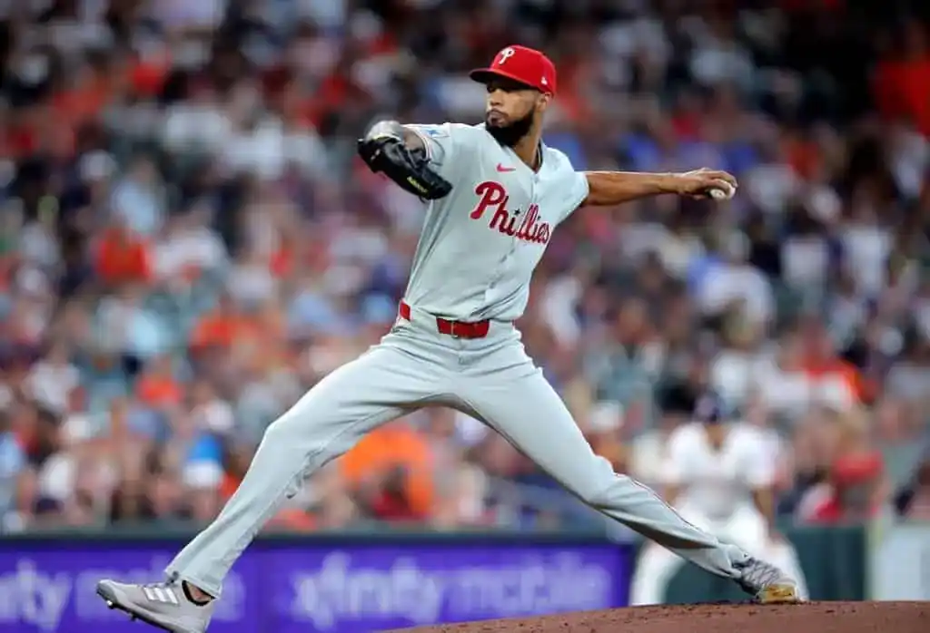 A Philadelphia Phillies pitcher throws a pitch on the baseball mound, wearing a gray uniform and a red cap, with fans in the background.