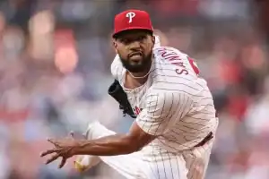 A baseball player in a striped jersey and red cap is in mid-pitch, showcasing dynamic movement on the field. Crowds and stadium lights in the background.