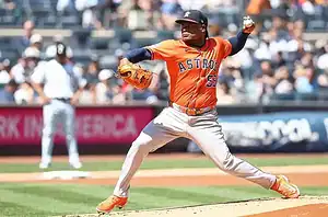 A Houston Astros pitcher in an orange jersey winds up to throw a pitch during a game.