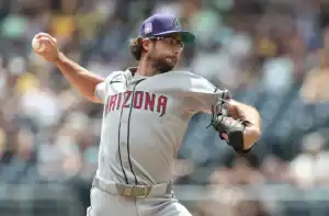 A pitcher wearing a gray Arizona Diamondbacks uniform throws a baseball in a sunny stadium, surrounded by blurred fans in the background.