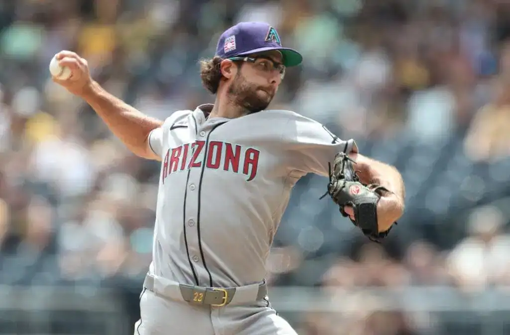 A pitcher wearing a gray Arizona Diamondbacks uniform throws a baseball in a sunny stadium, surrounded by blurred fans in the background.