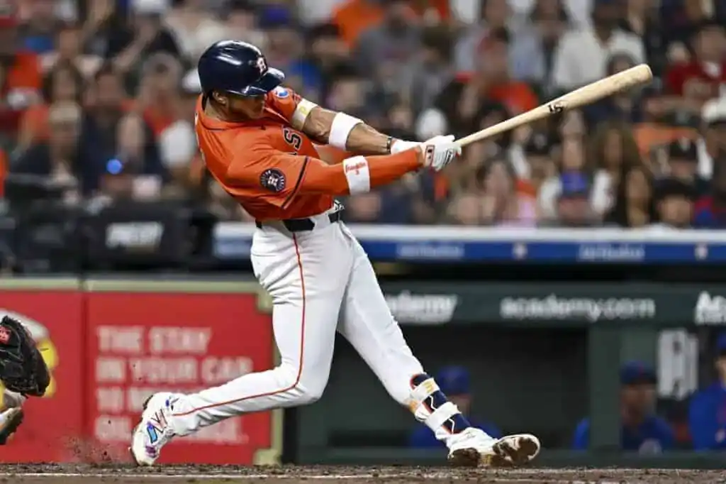 A baseball player in an orange uniform swings a bat, with a cheering crowd visible in the background.