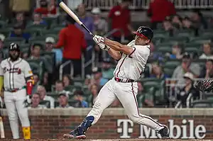 A baseball player in a white jersey swings a bat during a game, with a crowd visible in the background at a stadium.