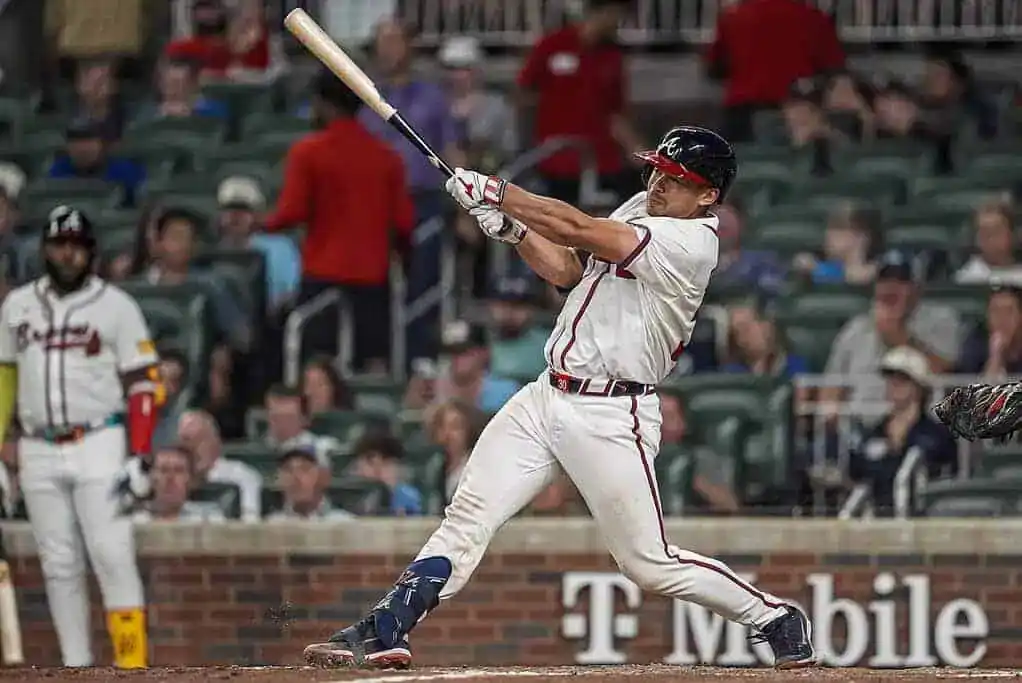 A baseball player in a white jersey swings a bat during a game, with a crowd visible in the background at a stadium.