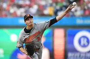 A pitcher in a gray Baltimore Orioles uniform winds up to throw a baseball during a game, surrounded by a colorful stadium crowd.