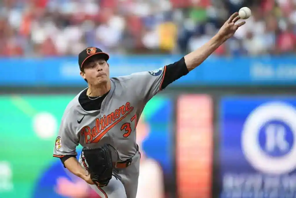 A pitcher in a gray Baltimore Orioles uniform winds up to throw a baseball during a game, surrounded by a colorful stadium crowd.