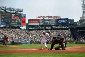 A baseball player swings at a pitch at Fenway Park, with a packed crowd and digital scoreboard in the background, showcasing statistics.