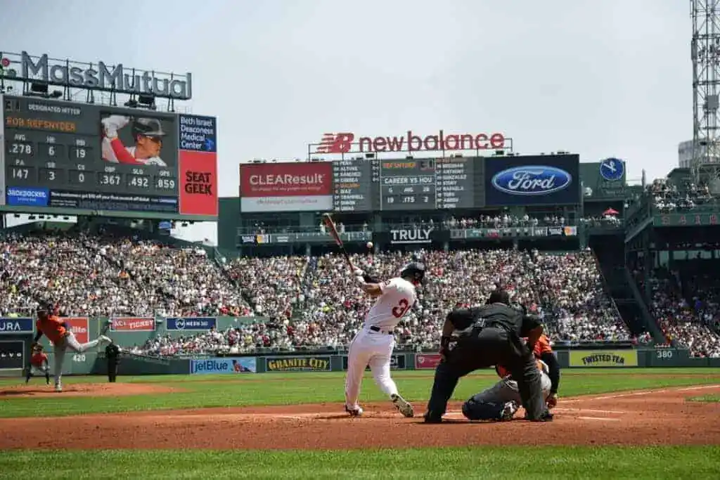 A baseball player swings at a pitch at Fenway Park, with a packed crowd and digital scoreboard in the background, showcasing statistics.