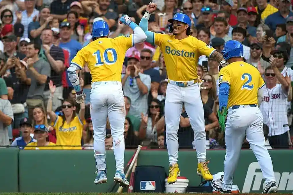 Boston Red Sox players in bright yellow City Connect uniforms celebrate a big play with high-fives as fans cheer in the background.