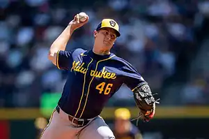 A Milwaukee Brewers pitcher throws a baseball, wearing a dark blue jersey with yellow accents and a grey cap during a sunny game.