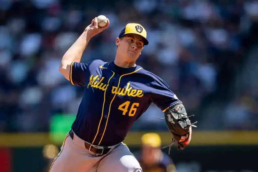 A Milwaukee Brewers pitcher throws a baseball, wearing a dark blue jersey with yellow accents and a grey cap during a sunny game.