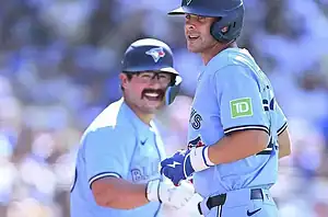 Two baseball players in light blue jerseys celebrate a play during a game, with a blurred crowd in the background.