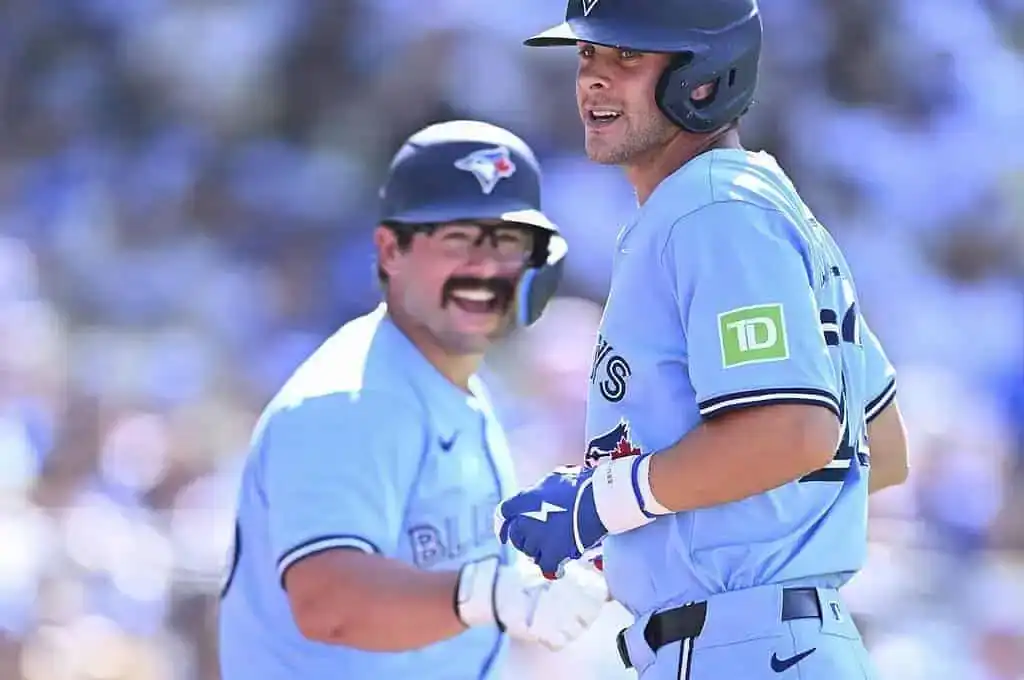 Two baseball players in light blue jerseys celebrate a play during a game, with a blurred crowd in the background.