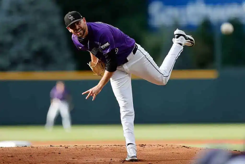 A baseball pitcher in a purple jersey winds up to throw the ball, showcasing an athletic stance and focus on the field.