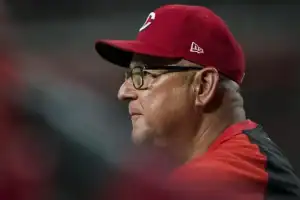 A coach wearing a red cap and jacket watches intently during a baseball game, with fans and players in the blurred background.