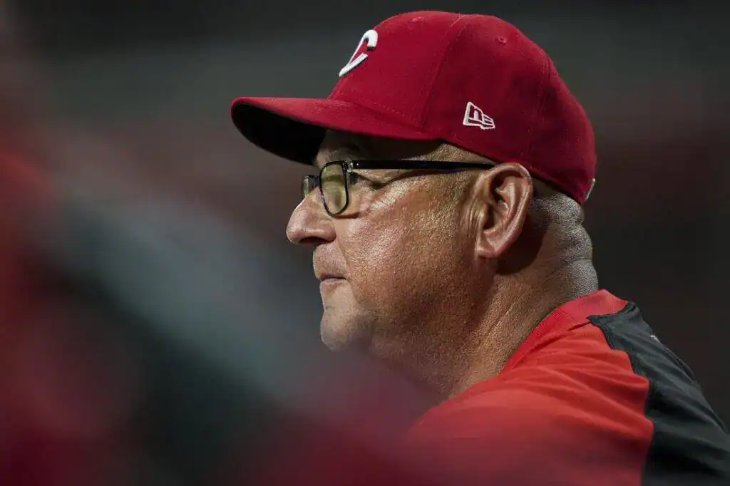 A coach wearing a red cap and jacket watches intently during a baseball game, with fans and players in the blurred background.