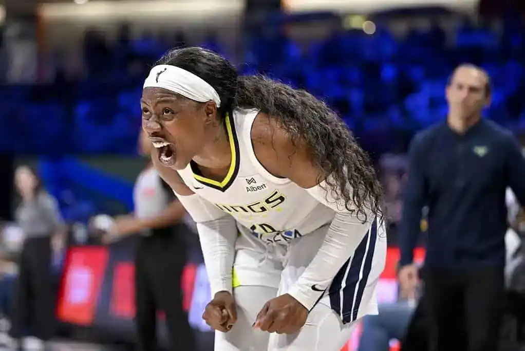 A basketball player in a white jersey with yellow and blue accents leans forward on the court, surrounded by blurred teammates and a vibrant crowd.