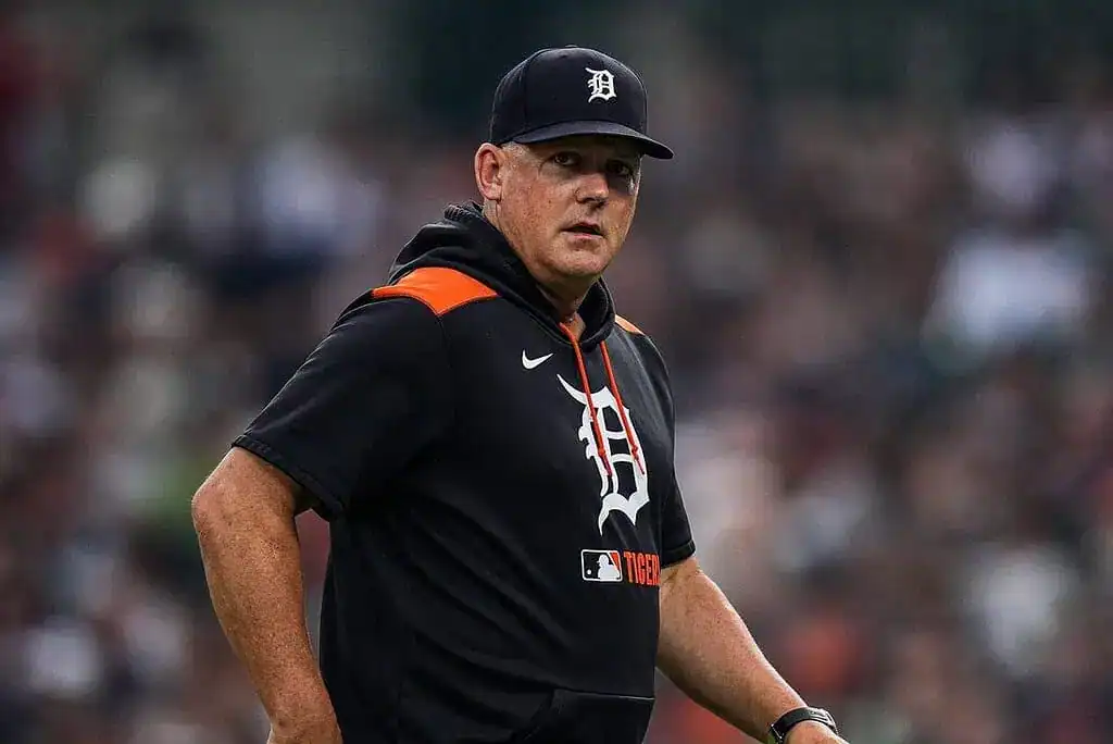 A man in a black and orange Detroit Tigers hoodie walks across a baseball field, wearing a cap with the Tigers logo.