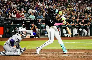 A baseball player swings a bat during a game, with a catcher crouching behind home plate and spectators in the background.