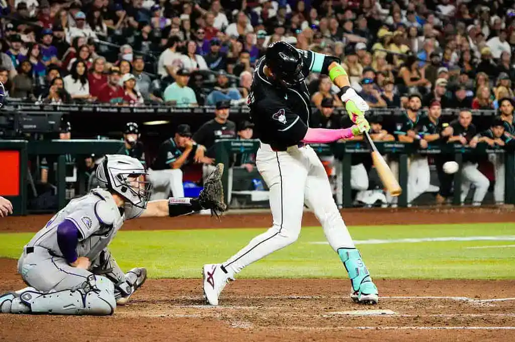 A baseball player swings a bat during a game, with a catcher crouching behind home plate and spectators in the background.