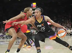 A basketball player in a black jersey dribbles the ball, evading a defender in a red jersey during an intense game action.
