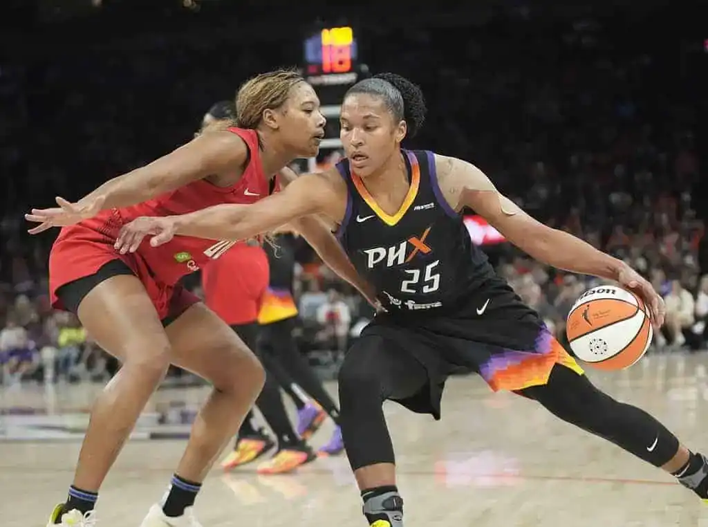 A basketball player in a black jersey dribbles the ball, evading a defender in a red jersey during an intense game action.