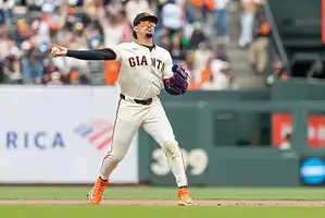 A San Francisco Giants pitcher in a white uniform and orange cleats prepares to throw a baseball on the field, with fans in the background.