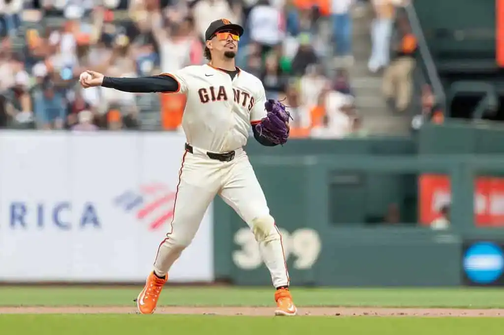 A San Francisco Giants pitcher in a white uniform and orange cleats prepares to throw a baseball on the field, with fans in the background.
